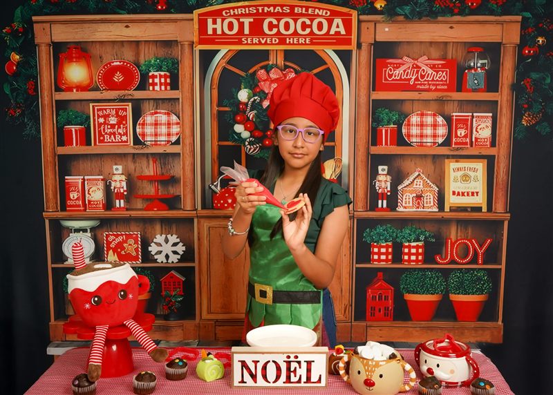 Niña con lentes y gorro de cocinero rojo decorando una galleta navideña