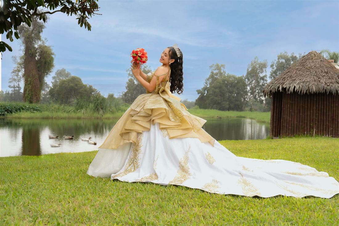 Quinceañera con vestido blanco y dorado junto a un lago, mirando su ramo de flores rojas