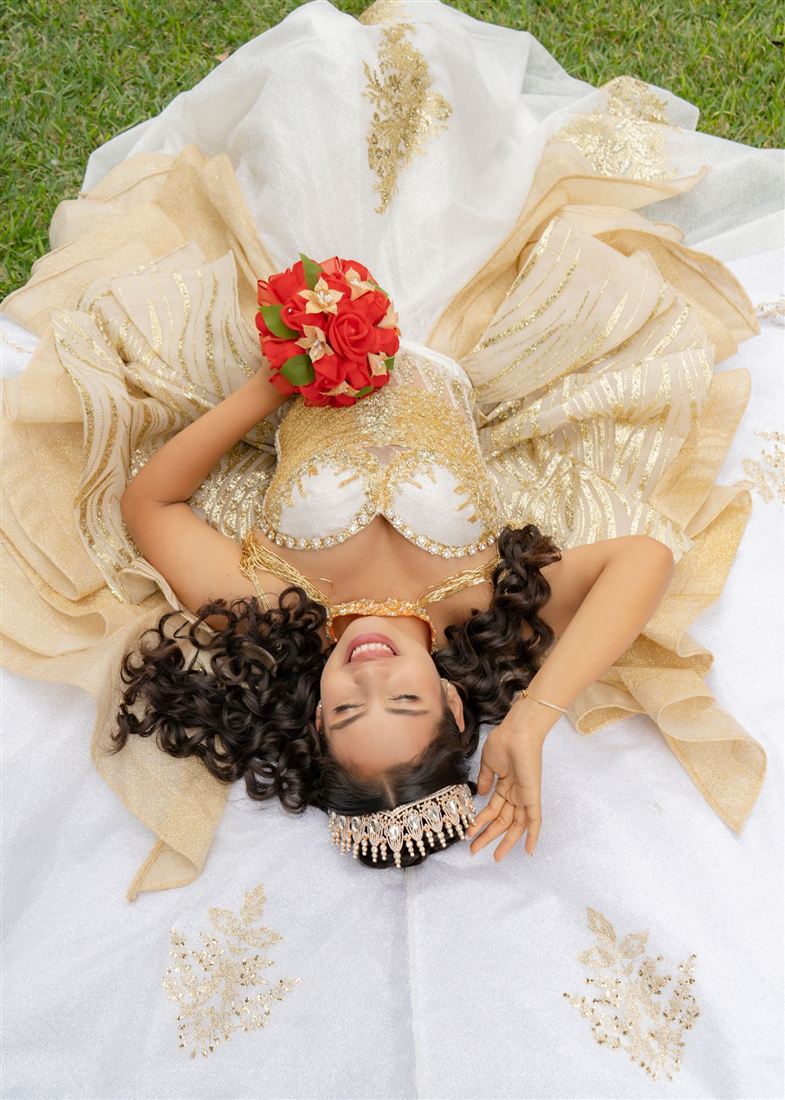 Quinceañera recostada sobre su vestido blanco y dorado con ramo de rosas rojas, vista desde arriba