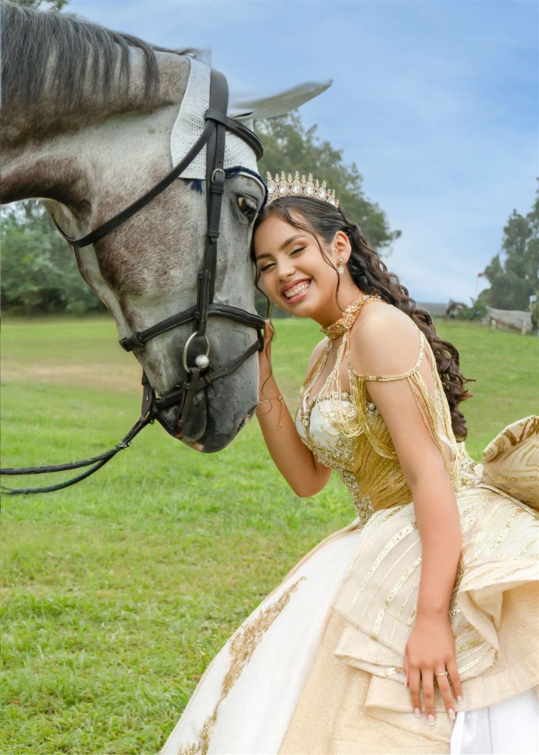 Quinceañera sonriente con corona apoyada en la cabeza de un caballo gris