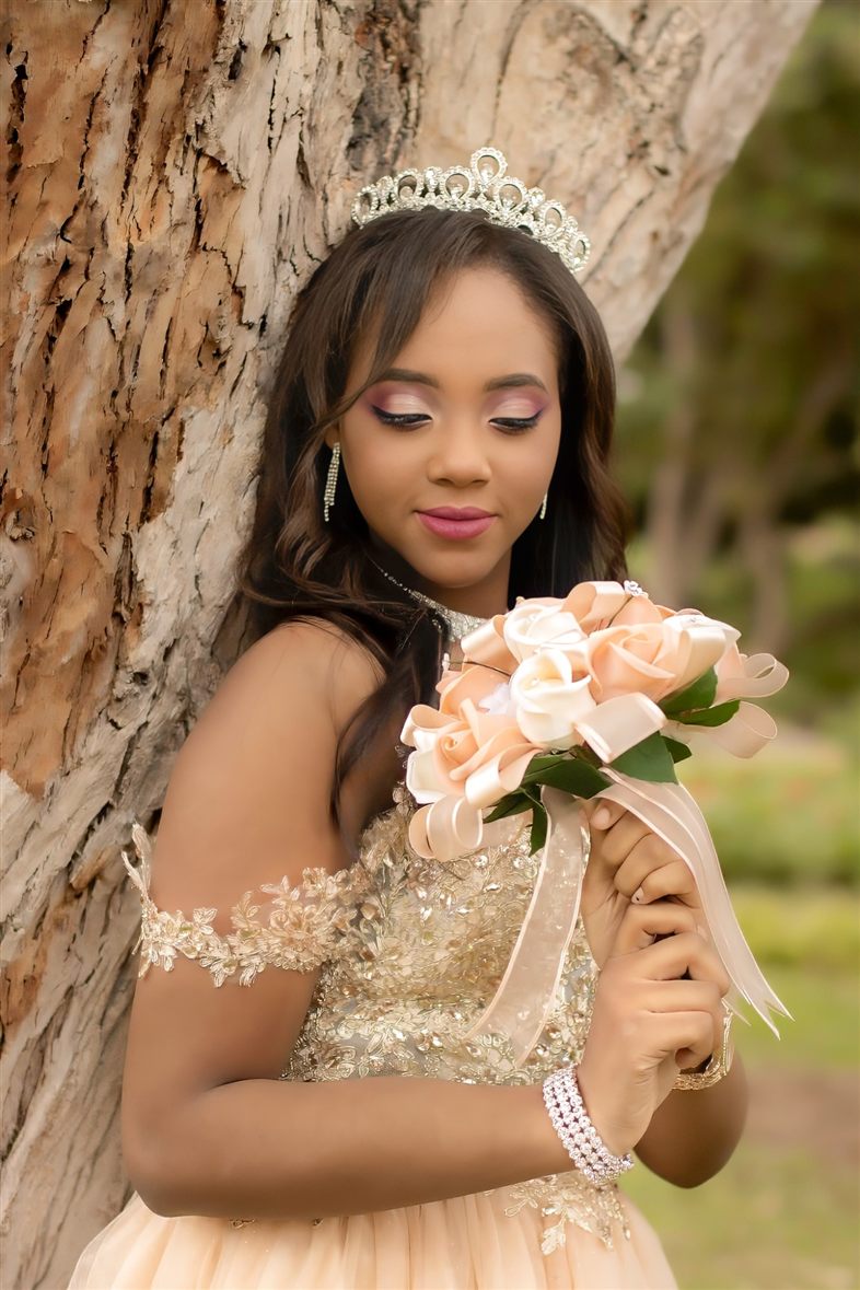 Quinceañera con tiara y vestido dorado apoyada en un tronco, mirando su ramo de rosas color durazno