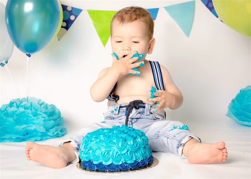 Bebé sentado comiendo su pastel azul con las manos, globos y pompones